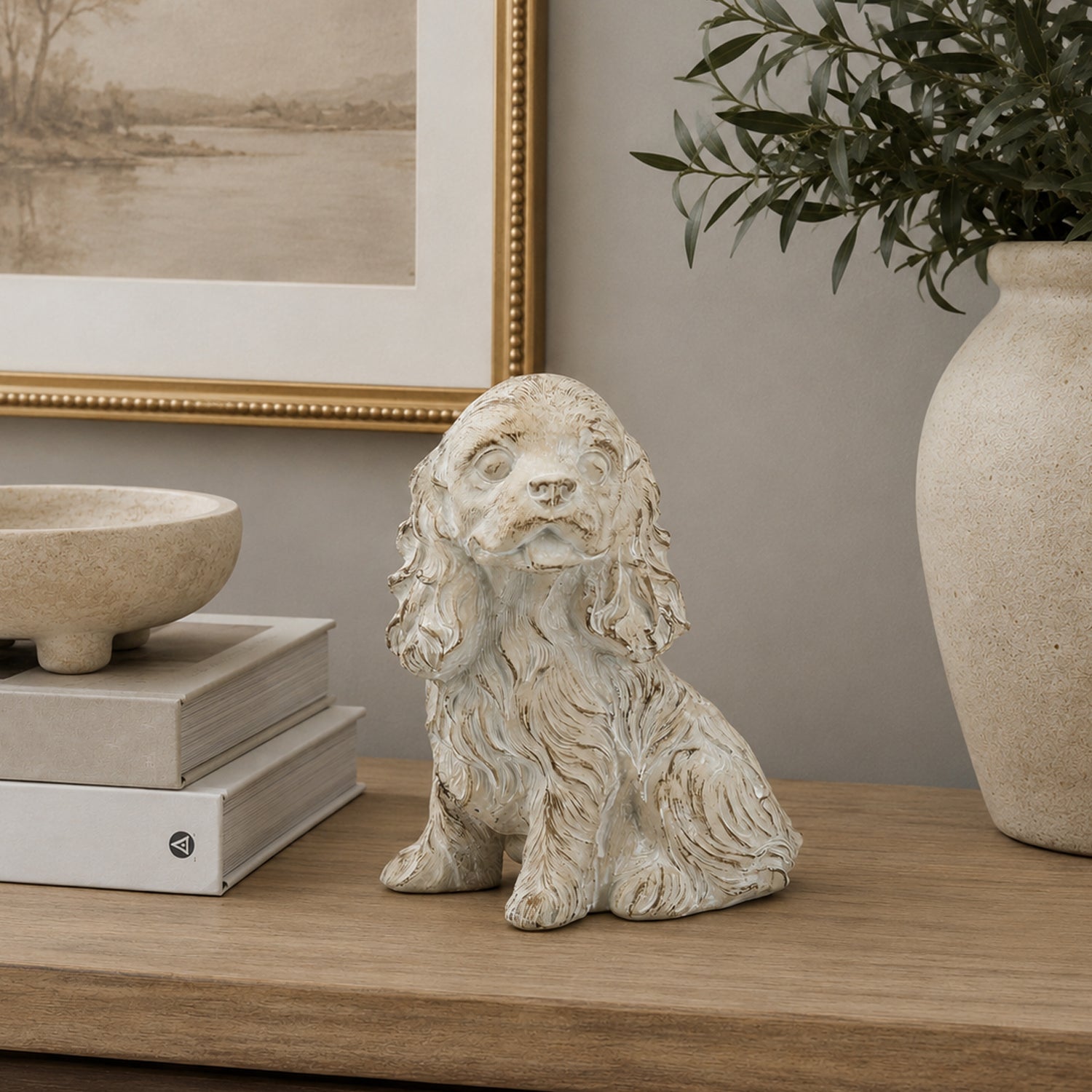 Design Republique Puppy Cement Statue in cream displayed on a wooden console with stacked books, framed artwork and decorative greenery.
