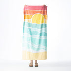 Multi-coloured beach towel featuring a sunset over aqua ocean waves, held up against a white background.