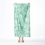 Mint green beach towel featuring an all-over tropical leaf print, held up against a white background.