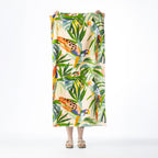 Multi-coloured Seaside Supplies beach towel featuring tropical birds and lush leafy foliage on a soft peach background, held up against a white background.