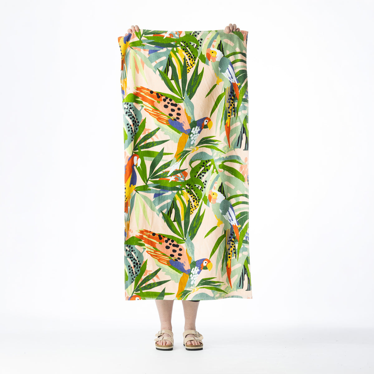 Multi-coloured Seaside Supplies beach towel featuring tropical birds and lush leafy foliage on a soft peach background, held up against a white background.