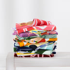 Colourful stack of assorted Seaside Supplies beach towels folded on a white bench with sunglasses on top.