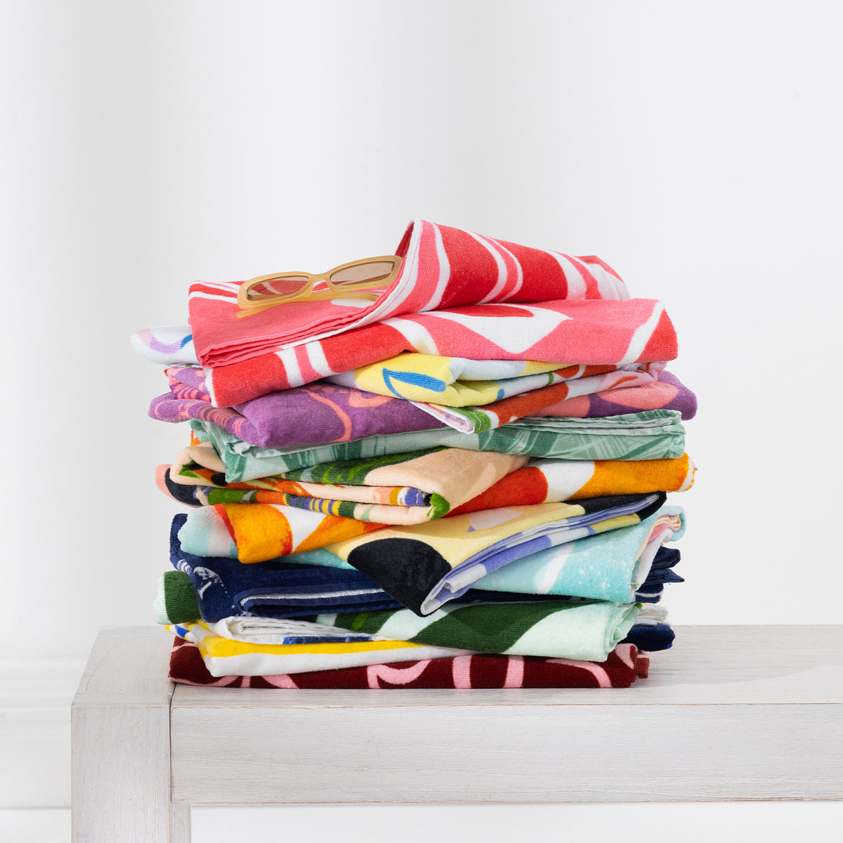 Colourful stack of assorted Seaside Supplies beach towels folded on a white bench with sunglasses on top.