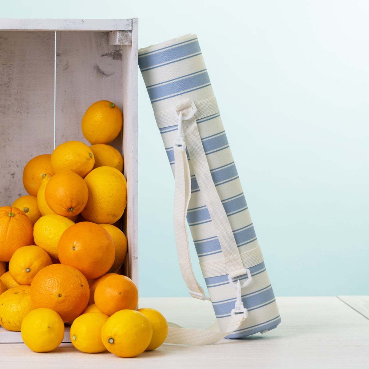 A picnic mat rolled up with a white shoulder strap stands next to a white crate filled with oranges on a light surface against a pale blue background.