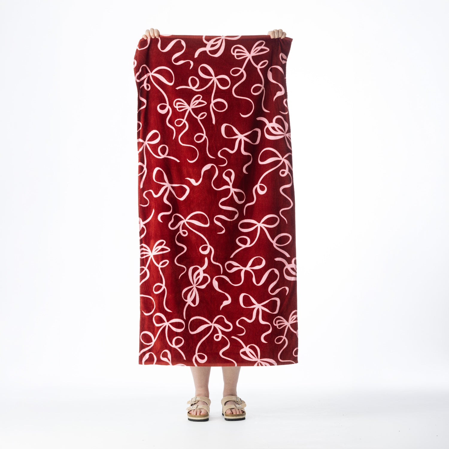 Deep red Seaside Supplies beach towel featuring an all-over pale pink bow and ribbon design, held up against a white background.