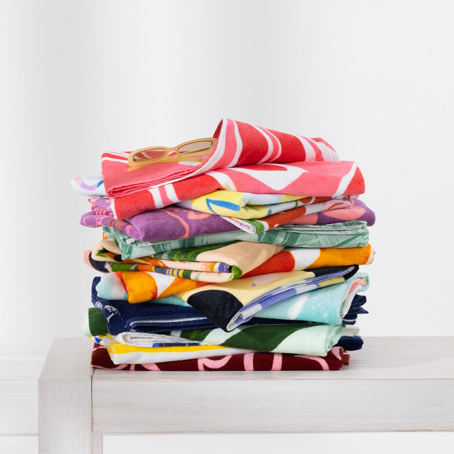 Colourful stack of assorted Seaside Supplies beach towels folded on a white bench with sunglasses on top.