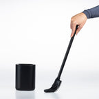 A black long-handled cleaning toilet brush  being held by a person's hand next to a matching black cylindrical holder on a clean white background.