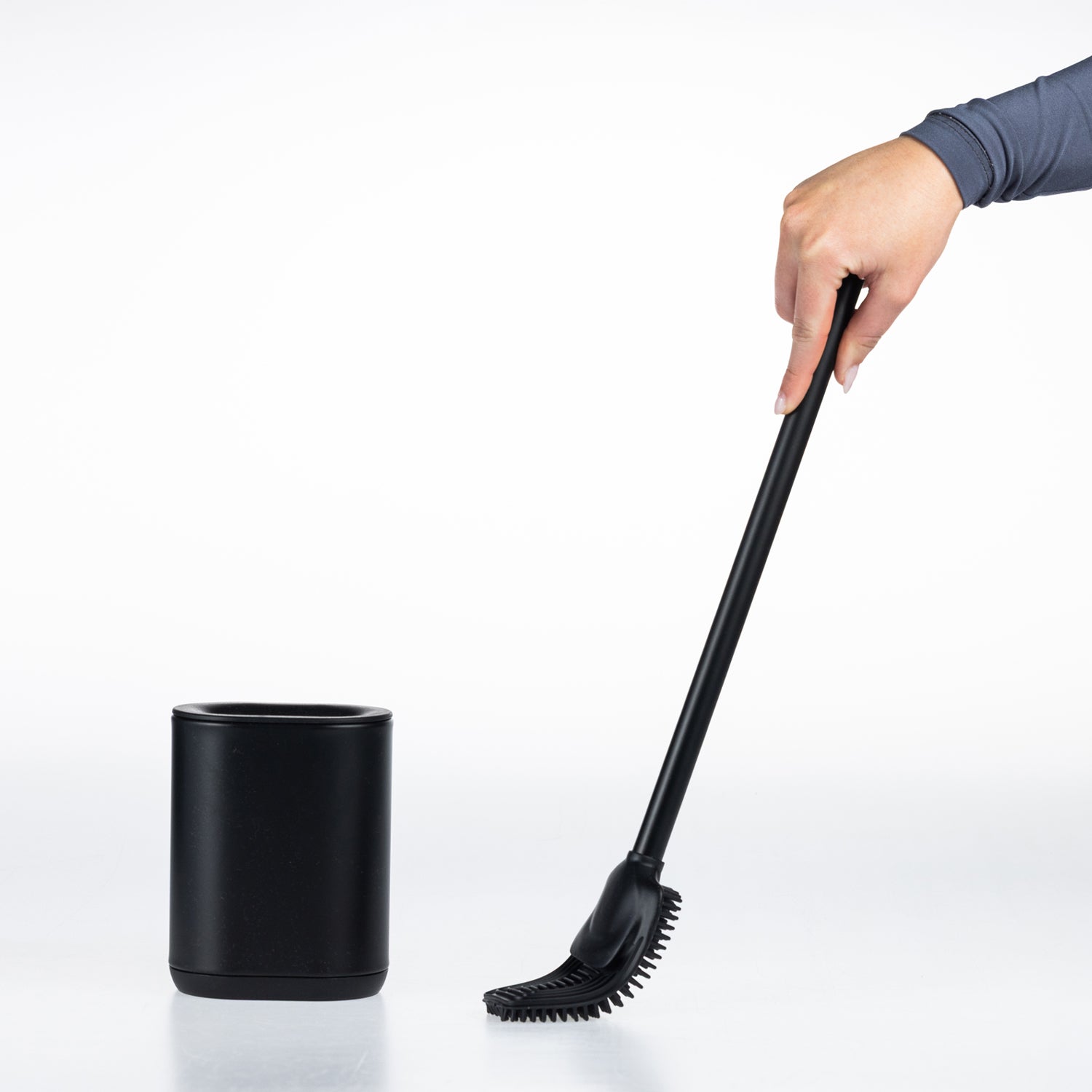 A black long-handled cleaning toilet brush  being held by a person's hand next to a matching black cylindrical holder on a clean white background.