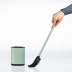 A hand holding a sage green and black toilet brush next to a matching cylindrical holder on a clean white background.