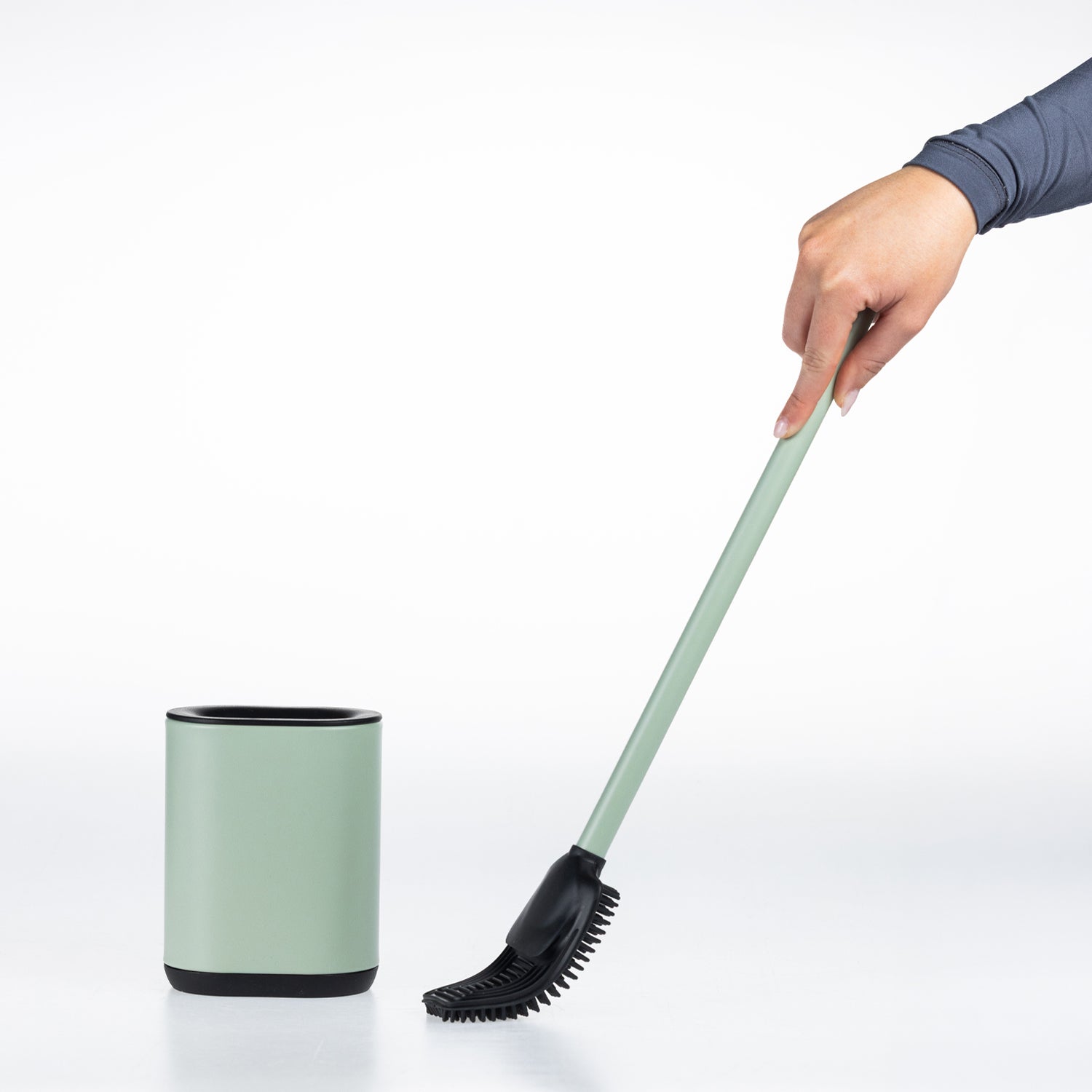 A hand holding a sage green and black toilet brush next to a matching cylindrical holder on a clean white background.