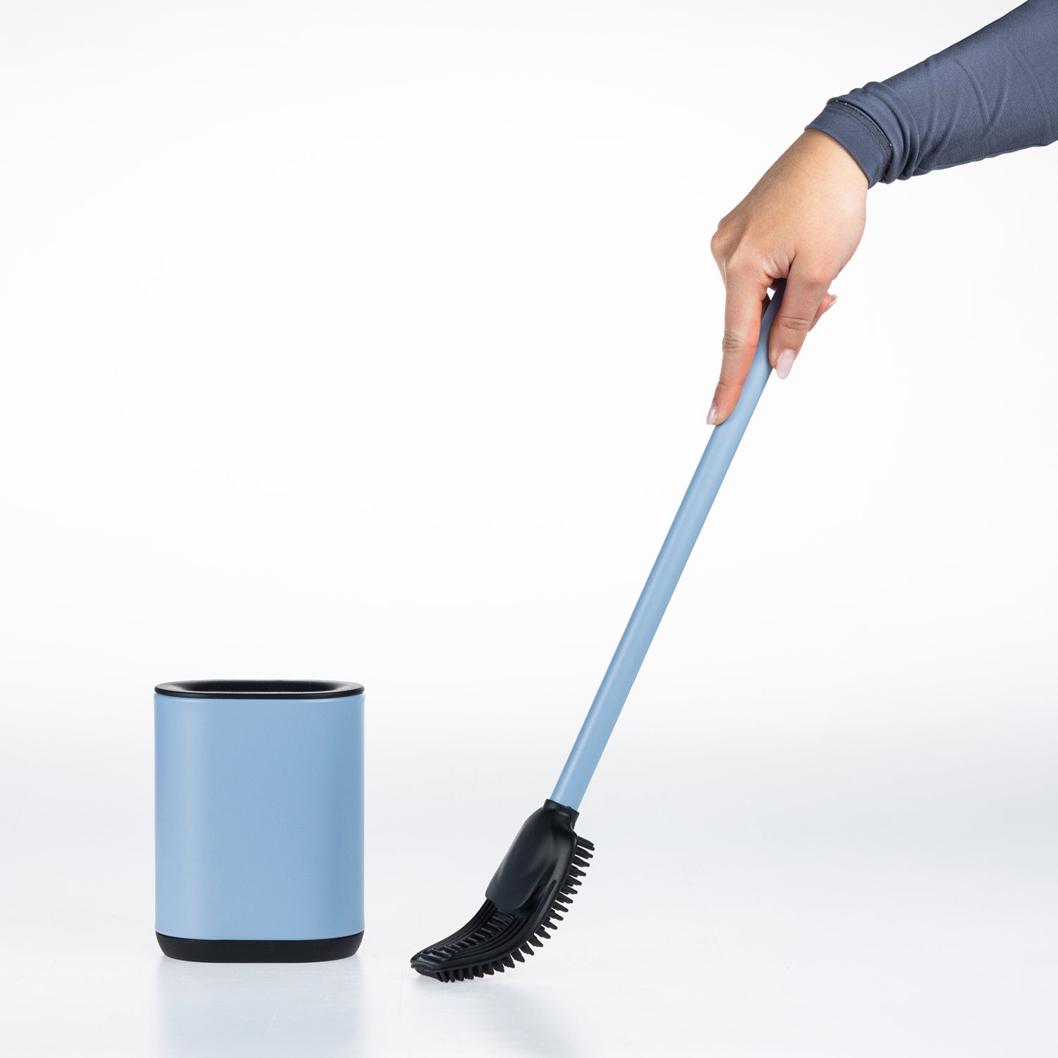 A hand holding a denim blue and black long-handled toilet brush next to a matching denim blue cylindrical container with black trim, set against a clean white background.