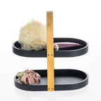 Black two-tier bathroom storage caddy with a bamboo carry handle, styled with a bath sponge, satin scrunchies and a cosmetic bottle.