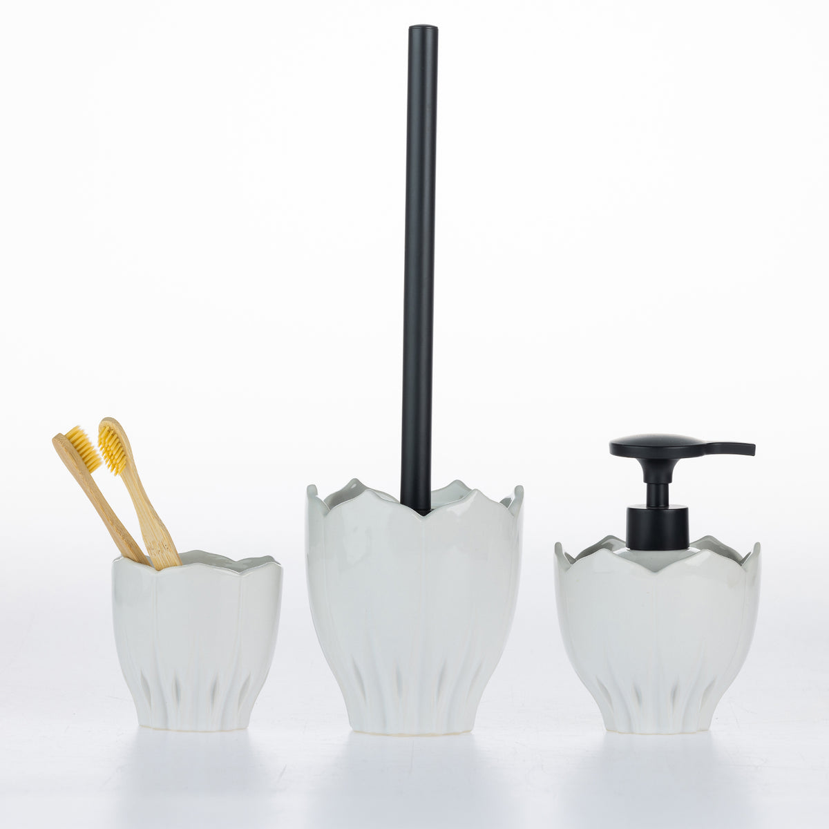 Three-piece ceramic bathroom set with a white flower-shaped toothbrush holder containing two bamboo toothbrushes, a matching toilet brush holder with a black handle, and a white flower-shaped pump dispenser with a black pump head, on a white background.