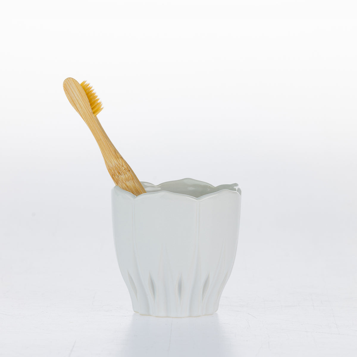 A white ceramic toothbrush holder with a fluted, scalloped rim, holding a light natural bamboo toothbrush, set against a seamless white background.