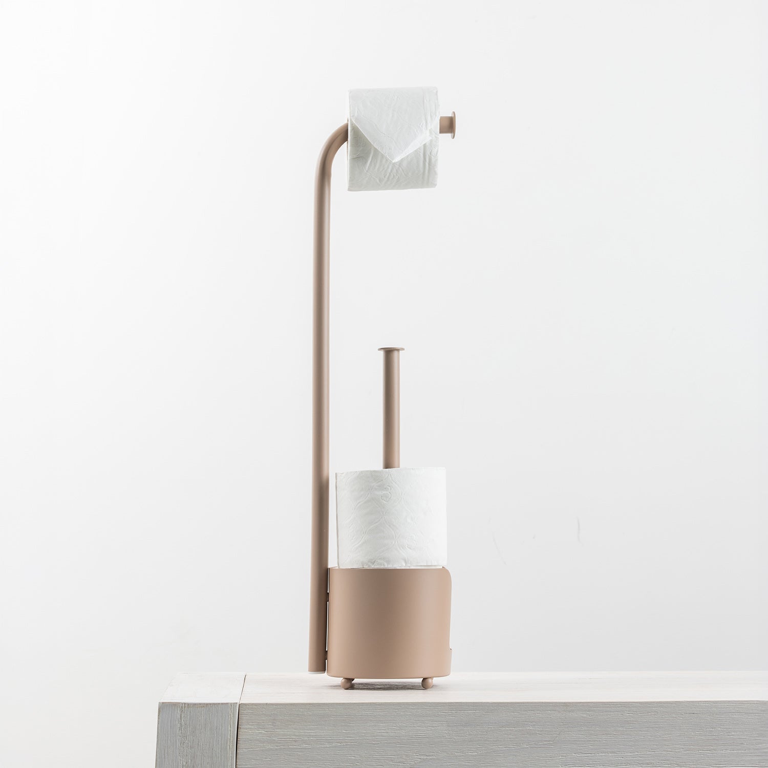 A modern toilet paper holder with a curved arm holding a roll of toilet paper, a cylindrical holder with a roll of paper, and a round base, shown on a light gray surface against a white background.
