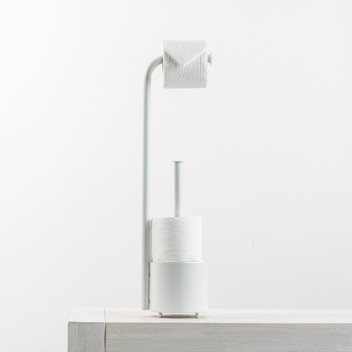 A minimalist white toilet paper holder and holder stand with two rolls of toilet paper, displayed on a light wood surface against a clean white background.