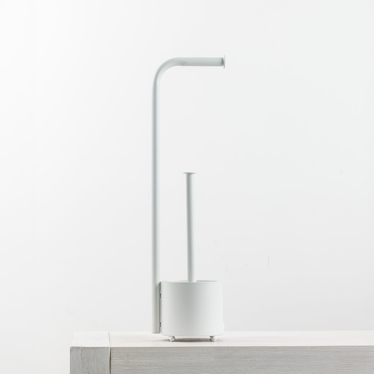 A minimalist white toilet paper holder with a curved arm and a cylindrical base, shown on a light wood surface against a white background.