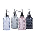 Four textured glass soap or lotion dispensers with silver pump bottles in light blue, gray, pink, and dark charcoal, arranged on a white background.