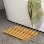 Bamboo bath mat styled beside a bathtub in a modern bathroom setting with soft green towel accents and terrazzo flooring.