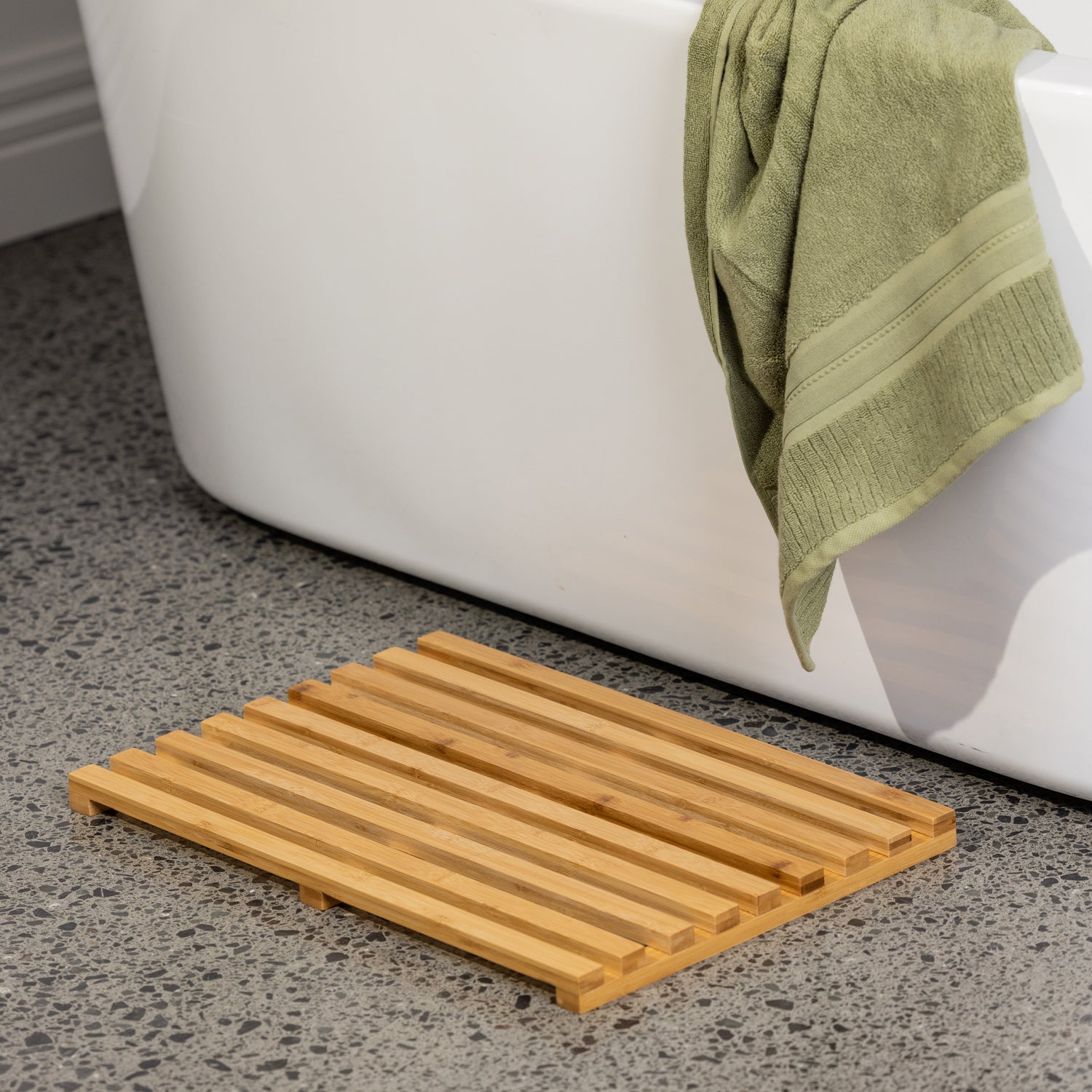 Bamboo bath mat styled beside a bathtub in a modern bathroom setting with soft green towel accents and terrazzo flooring.