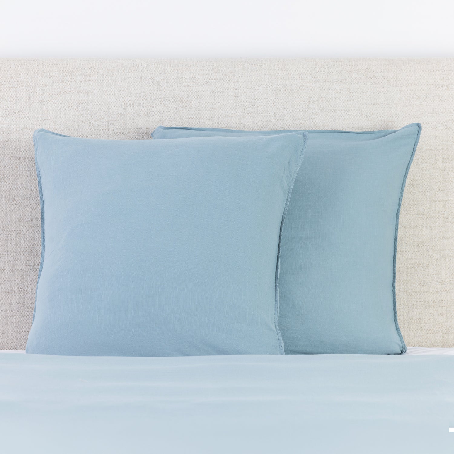 Dusty blue linen look European pillowcases styled on a bed with a neutral fabric headboard.