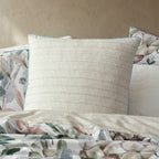 A European pillow with a cream cotton fabric and thin light blue horizontal stripes, styled on a bed with matching floral bedding and a textured beige wall background.