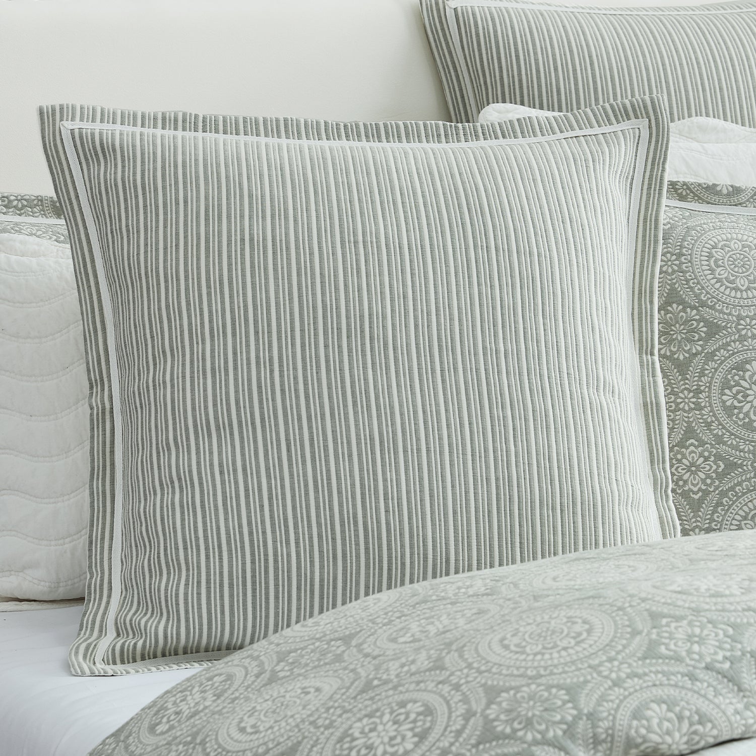 A square decorative throw pillow with vertical gray and white stripes, styled on a bed with matching patterned bedding and pillows.