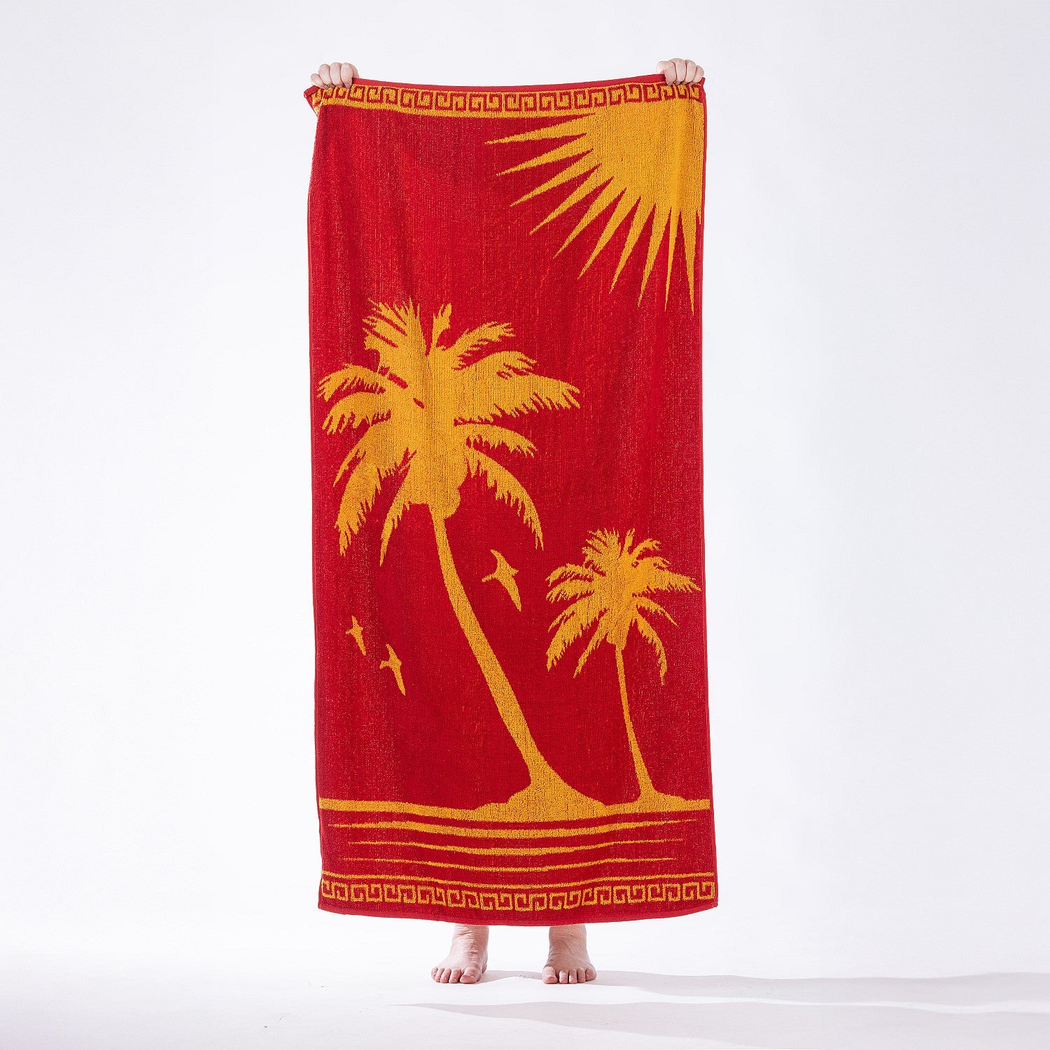 Bright tropical beach towel featuring palm tree silhouettes and sun motifs in bold red and yellow tones, held up against a plain studio backdrop.