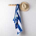 A blue and white striped beach towel hangs from a light wood wall hook, with a straw hat resting on the adjacent hook against a plain light gray wall.