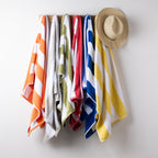 Six striped beach towels in orange, white, olive green, red, blue, and yellow hang from a wooden rack on a light gray wall, with a straw hat hanging on the right side.