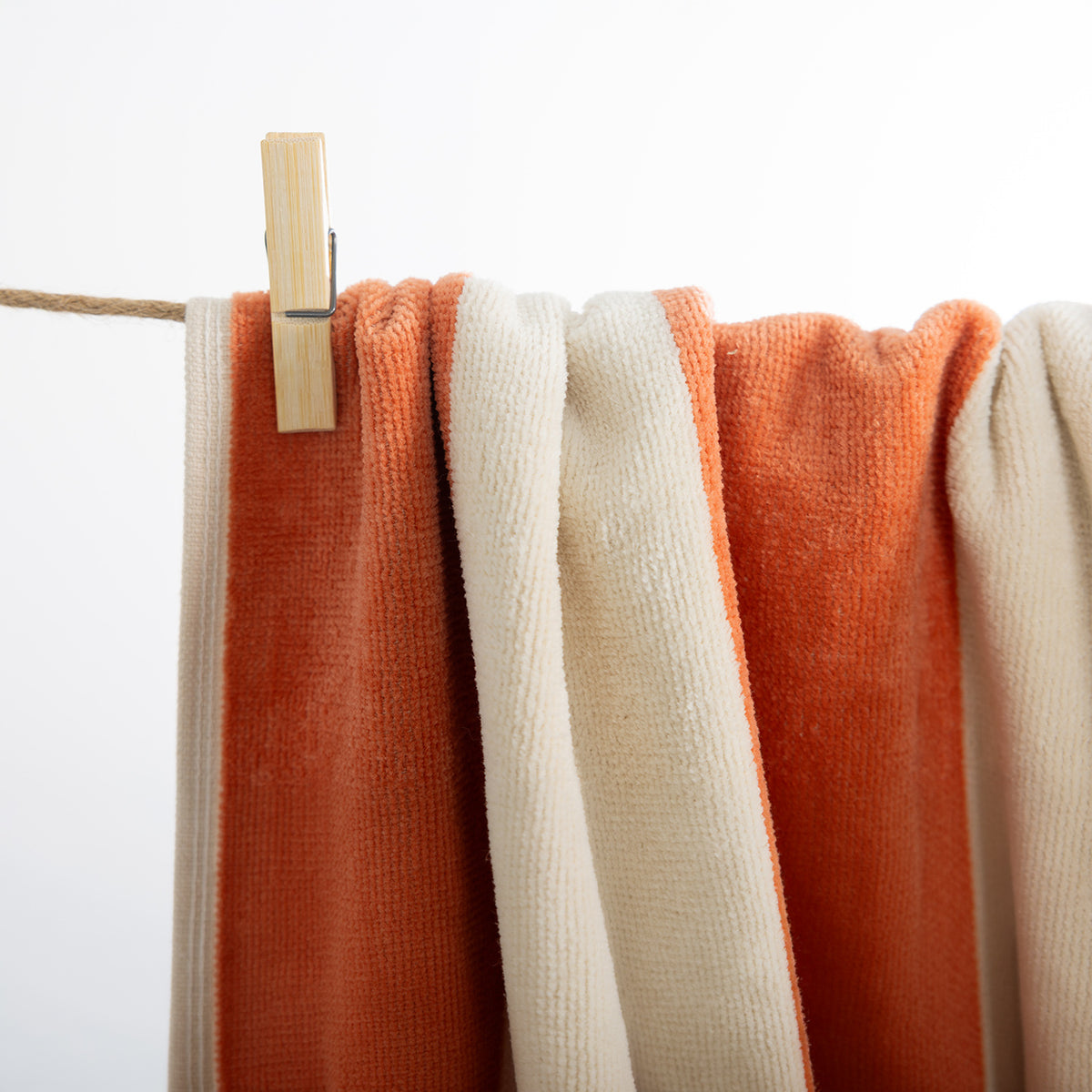 A close-up of two textured towels hanging from a rope with a wooden clothes clip, in orange and cream colors against a plain white background.