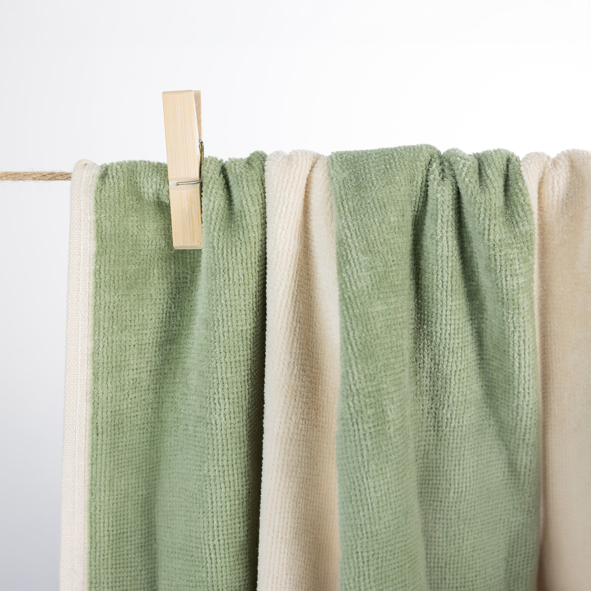 A close-up of soft textured fabric towels hanging from a rope line with a wooden clothes clip, shown in sage green and cream against a white background.