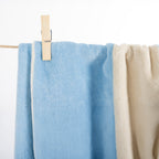 A light blue textured towel hanging from a rope line with a wooden clothes clip, next to a cream-colored towel against a white background.