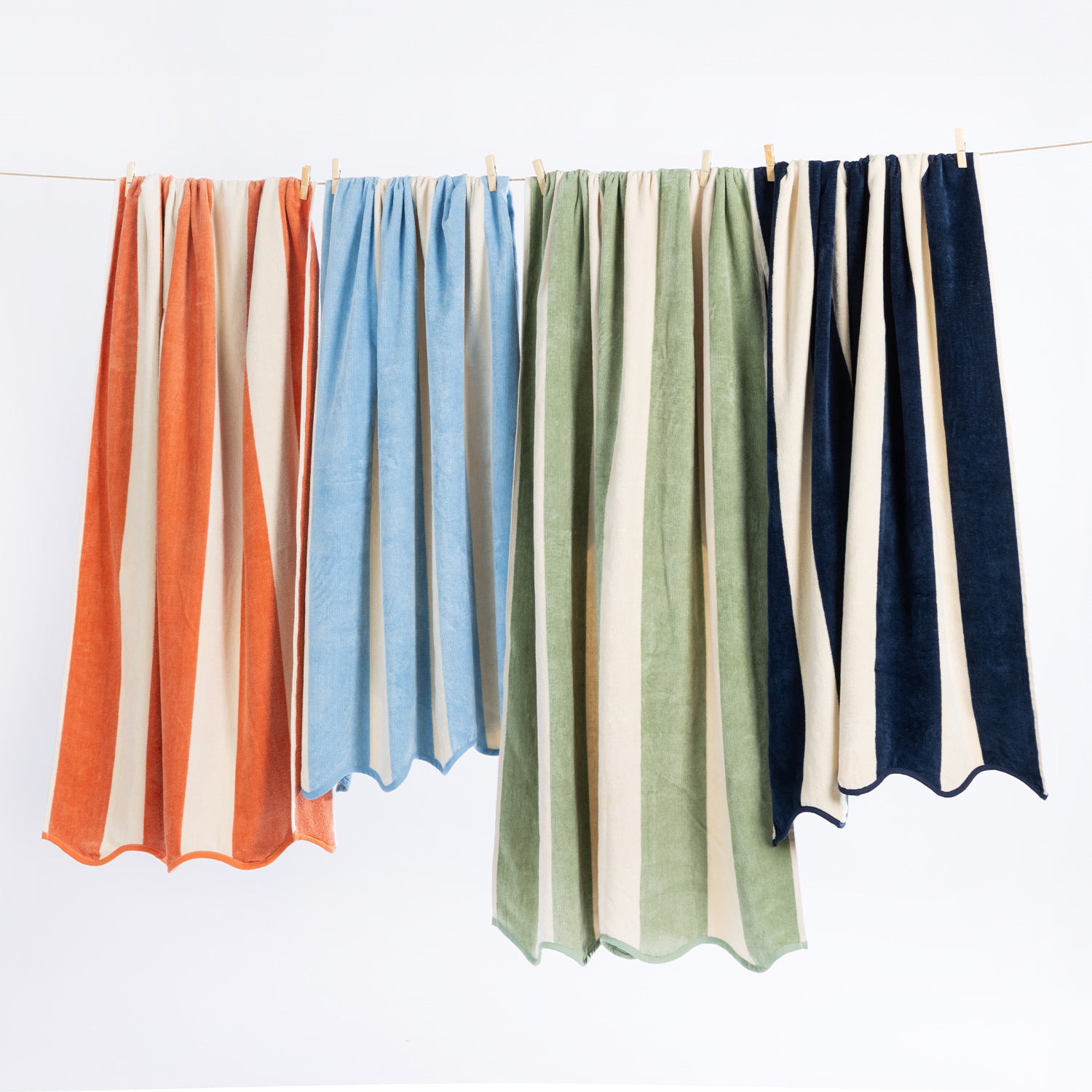 Four striped bath towels hanging from a clothesline with wooden clips against a plain white background. The towels feature vertical stripes in orange and cream, light blue and cream, sage green and cream, and navy blue and cream.