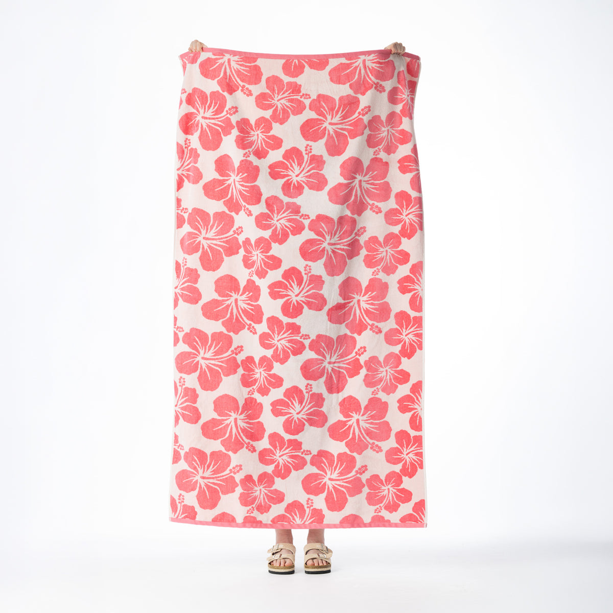 Pink beach towel featuring an all-over hibiscus floral pattern, held up against a white background.
