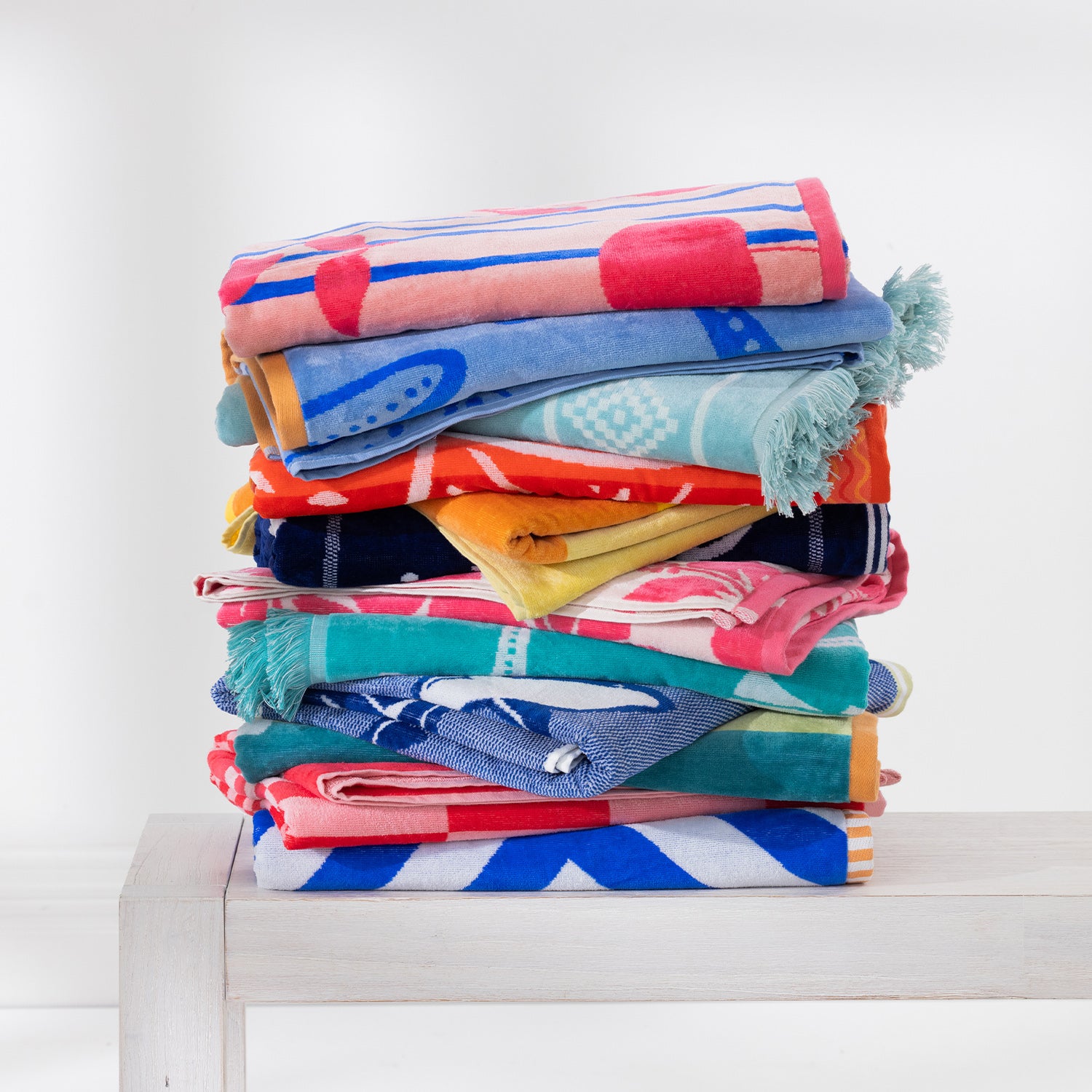 Stack of colourful patterned beach towels folded on a white bench against a white background.