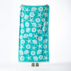 Aqua beach towel featuring an all-over seashell, starfish and sand dollar design with fringed edges, held up against a white background.