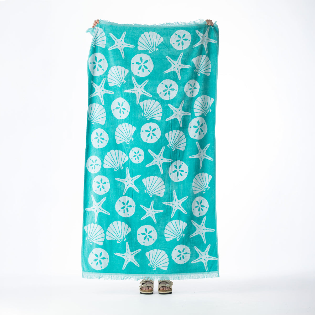 Aqua beach towel featuring an all-over seashell, starfish and sand dollar design with fringed edges, held up against a white background.