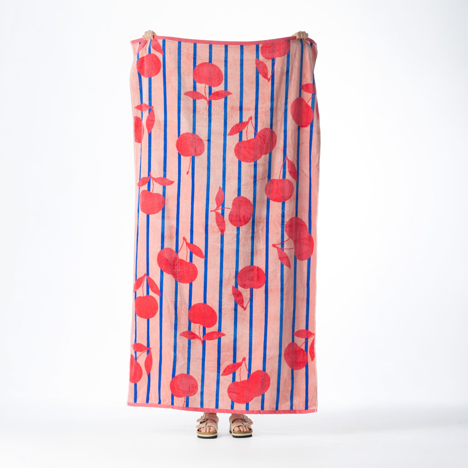 Pink beach towel featuring red cherries and blue vertical stripes, held up against a white background.