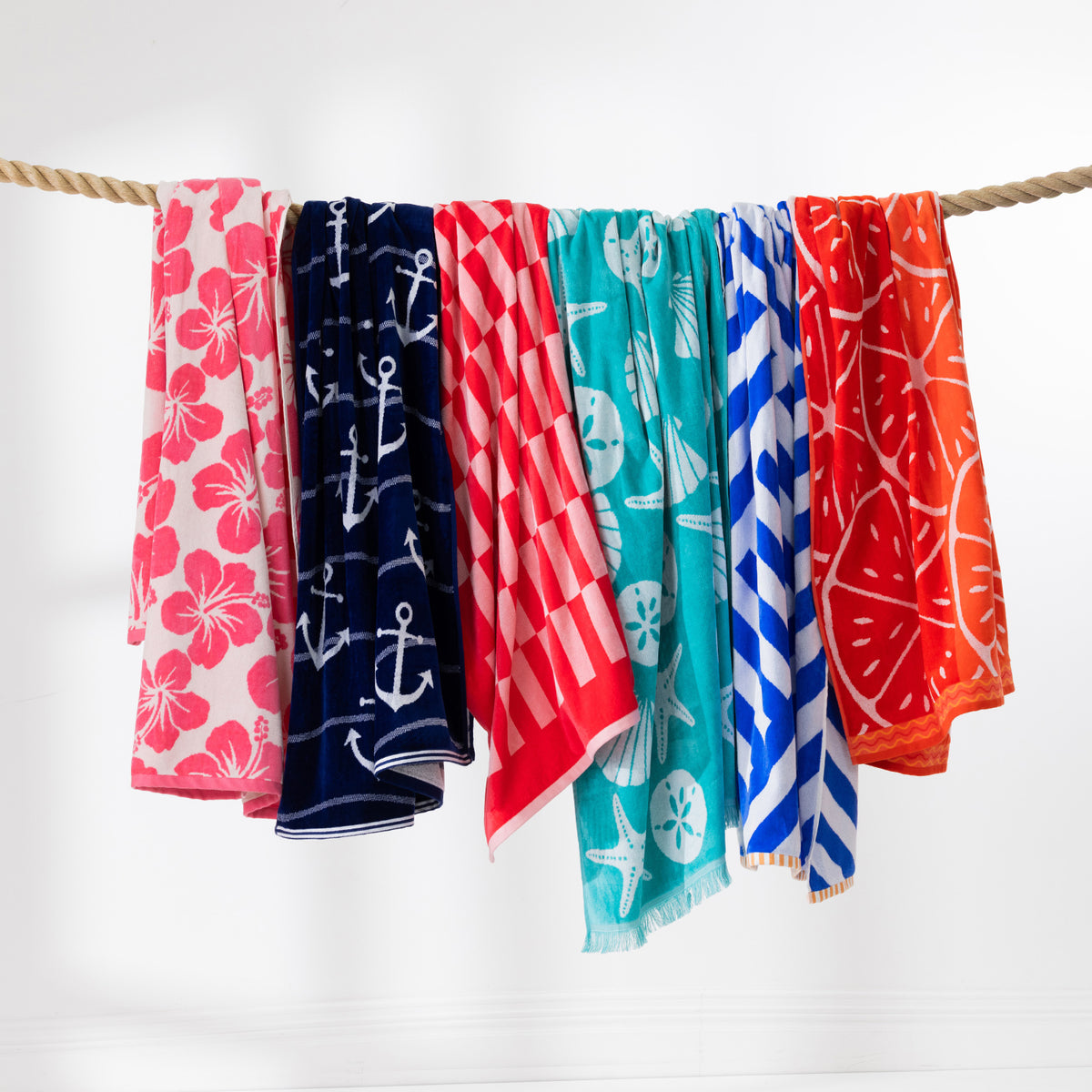 Colourful beach towel range draped over a rope, featuring floral, anchor, shell, striped and citrus designs.