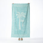 Eucalyptus beach towel featuring white palm trees, a geometric border and fringed edges, held up against a white background.