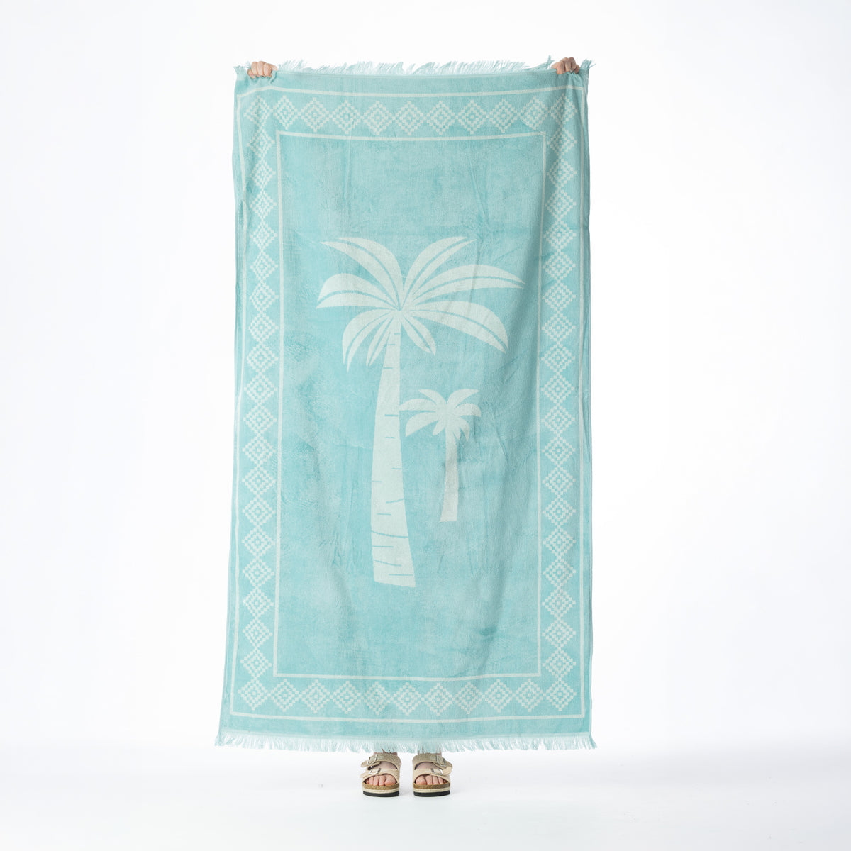 Eucalyptus beach towel featuring white palm trees, a geometric border and fringed edges, held up against a white background.