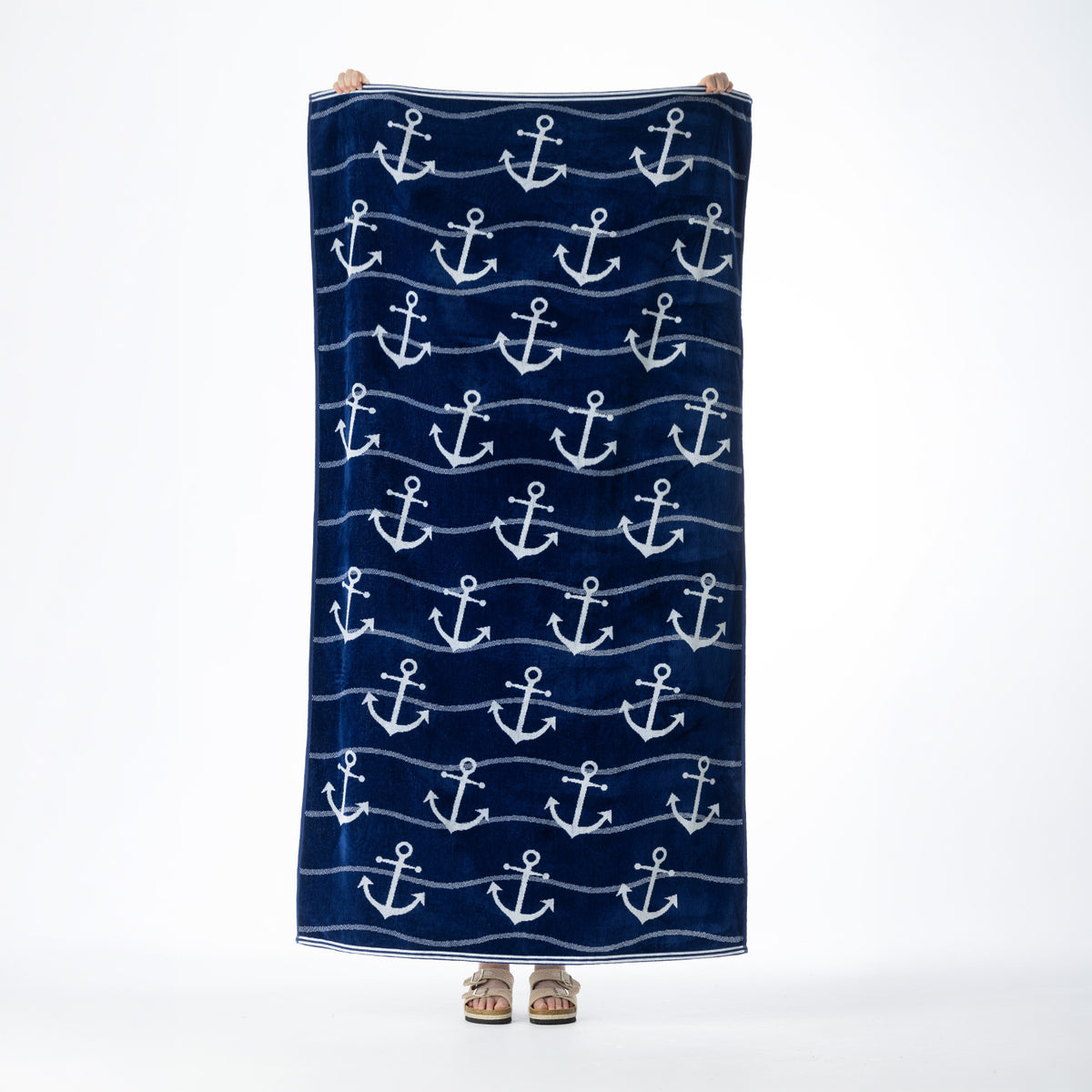 Navy blue beach towel featuring an all-over white anchor design with wavy stripe detailing, held up against a white background.