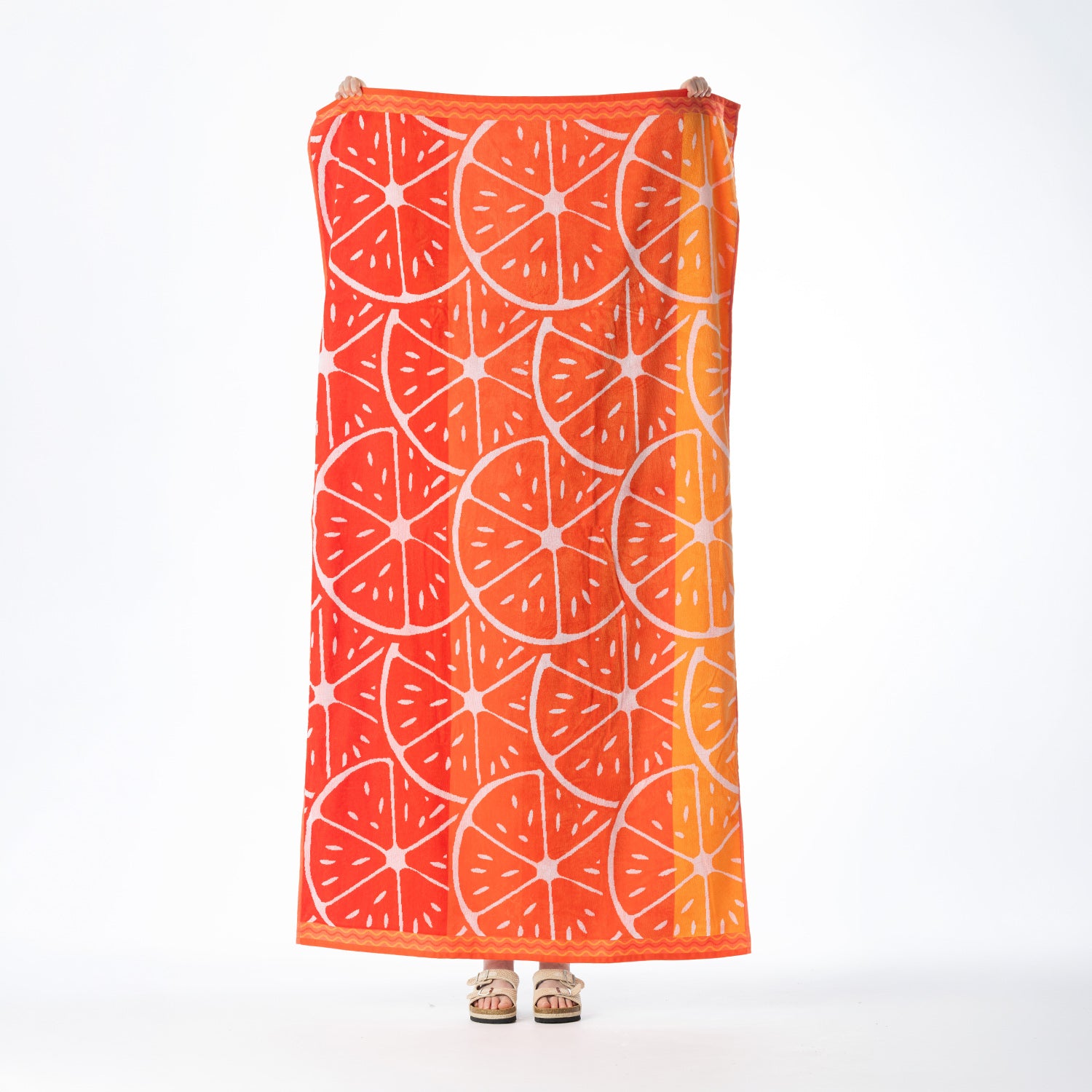 Orange beach towel featuring a bold citrus slice design with contrasting pale pink detailing and decorative borders, held up against a white background.