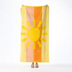 Yellow and orange beach towel featuring a large sunshine design over warm vertical stripes, held up against a white background.