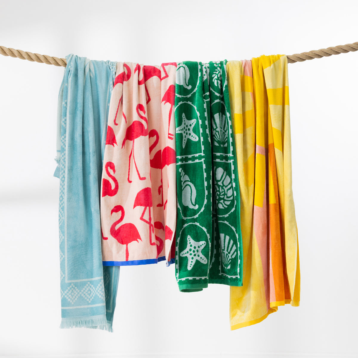 Colourful beach towels in assorted coastal and summer designs draped over a rope against a white background.