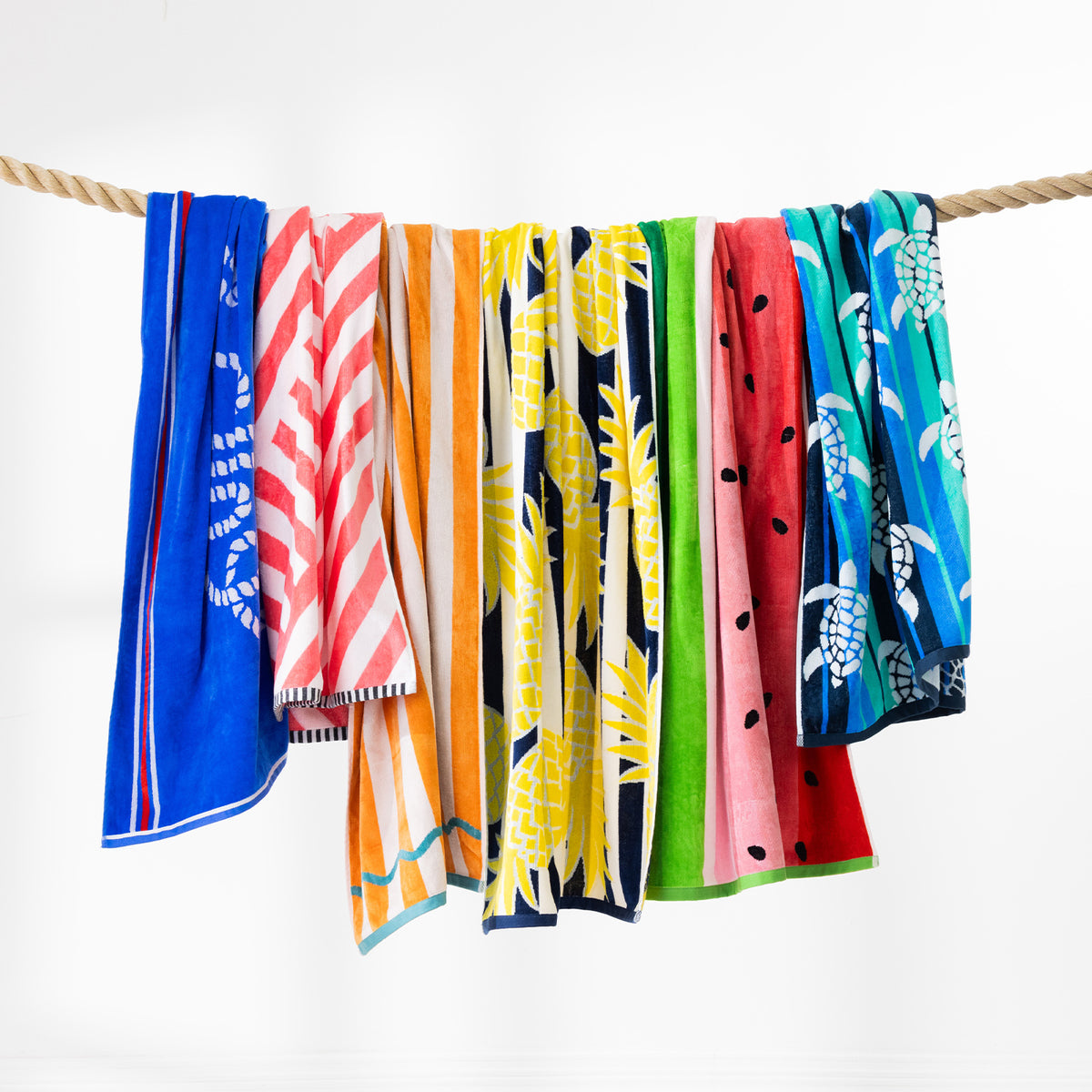 Colourful beach towels featuring assorted tropical and summer designs draped over a rope against a white background.