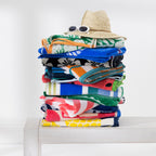 Stack of colourful folded beach towels topped with a straw sunhat and white sunglasses on a white wooden bench.