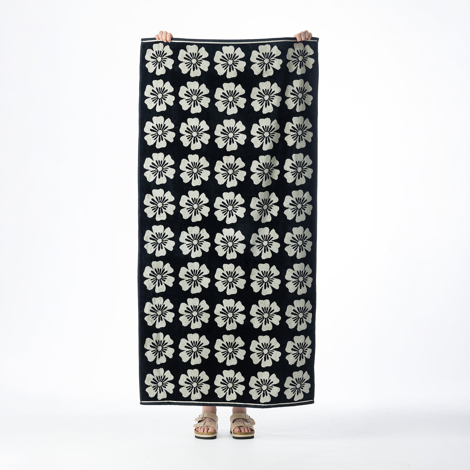 Black beach towel featuring a repeating cream floral design, held up against a white background.