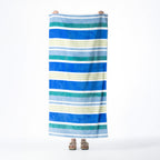 Multicoloured beach towel featuring horizontal stripes in blue, teal, light blue, white and pale green, held up against a white background.